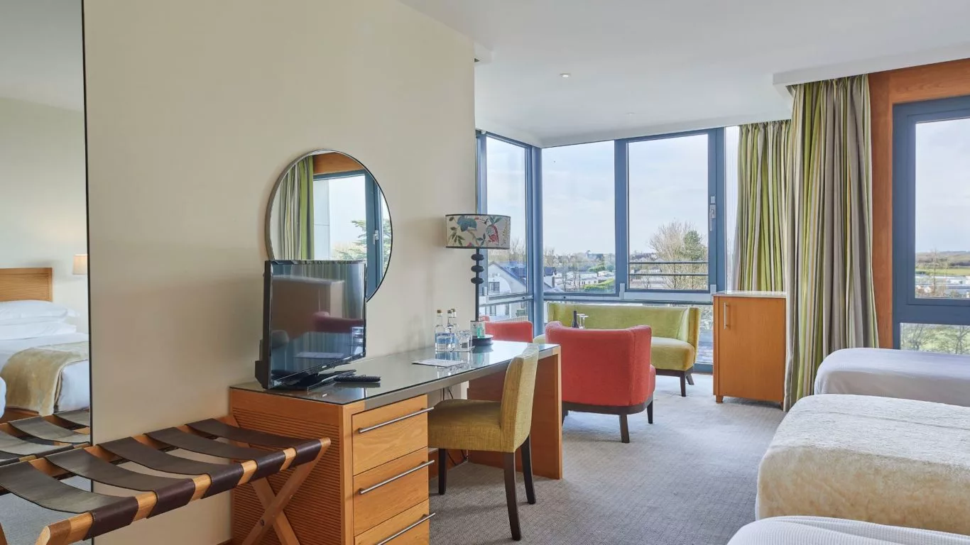 Spacious hotel room with cosy seating area, large windows with scenic views, desk, TV, and vibrant décor.