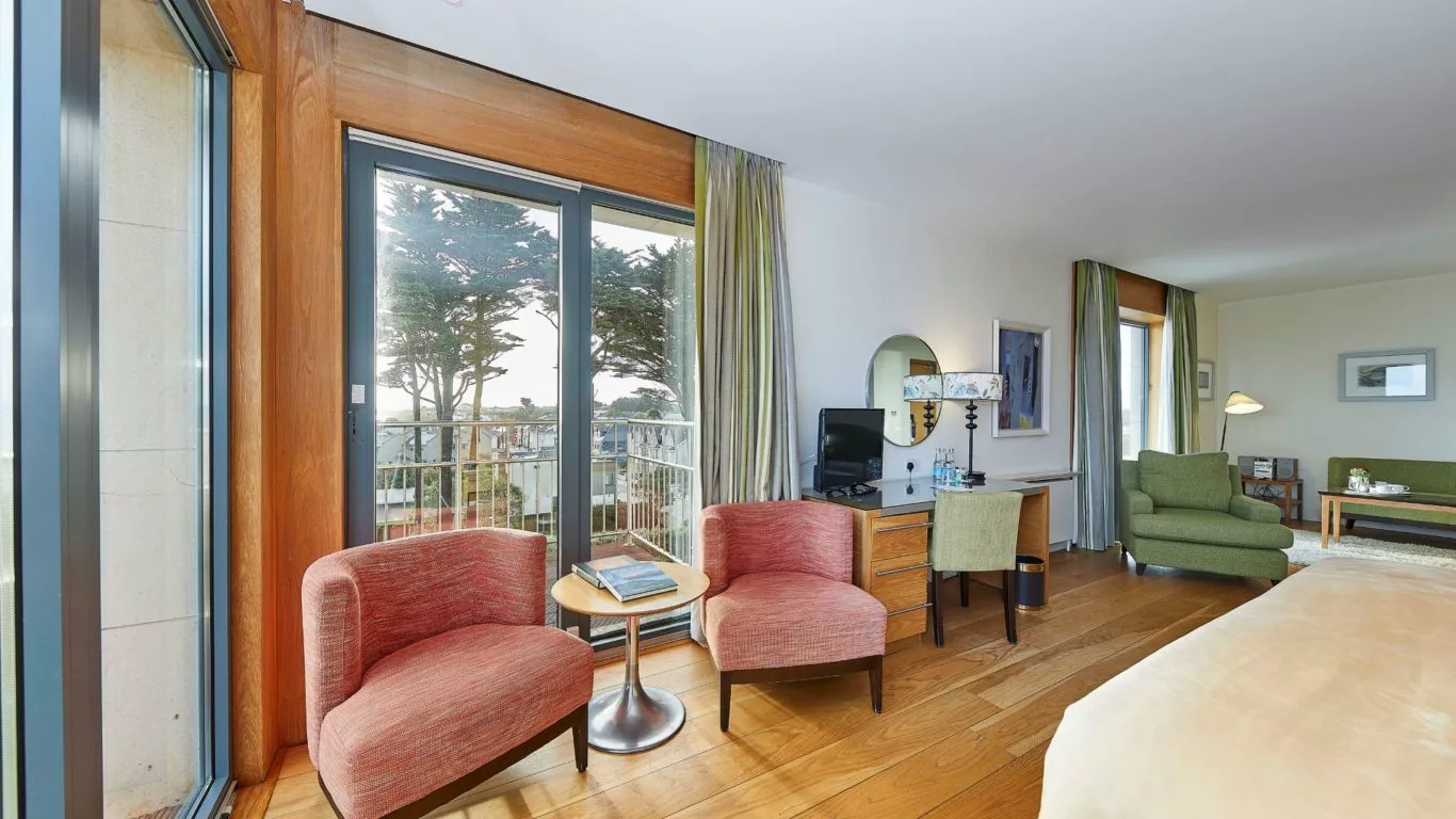 Spacious hotel room with balcony, cosy red chairs, modern decor, and sea view, promising a relaxing seaside escape.