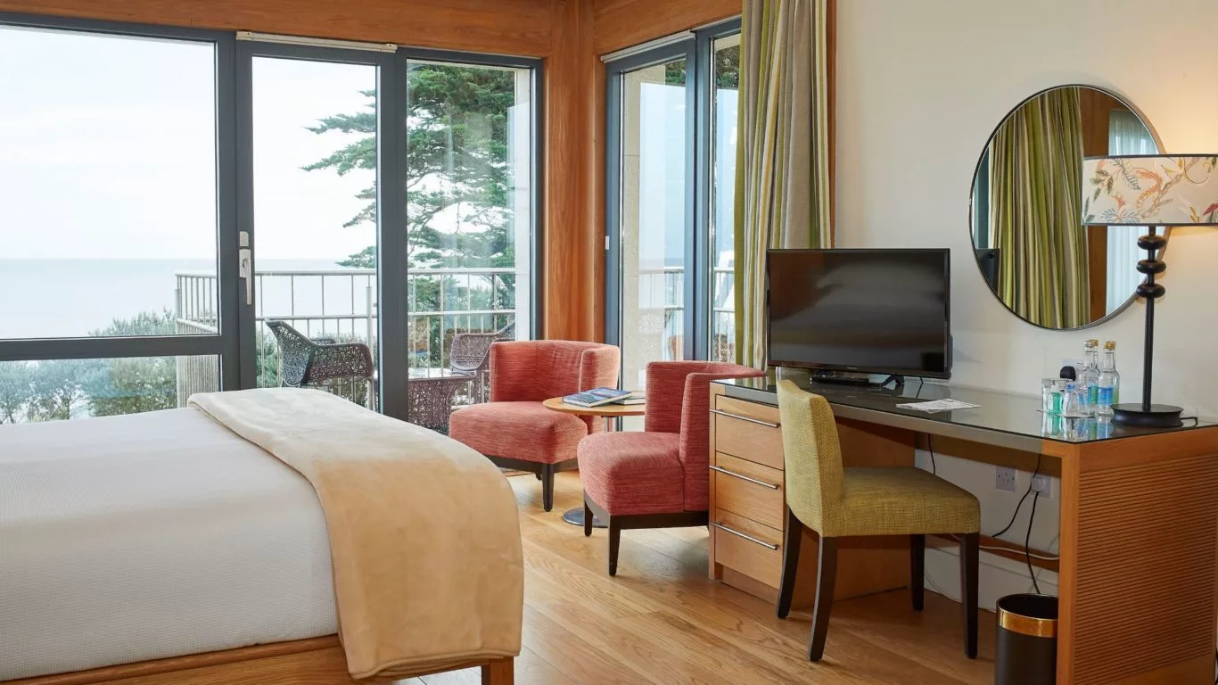 Spacious hotel room with sea view, balcony, elegant seating, flat-screen TV, and desk. Bright, relaxing atmosphere.