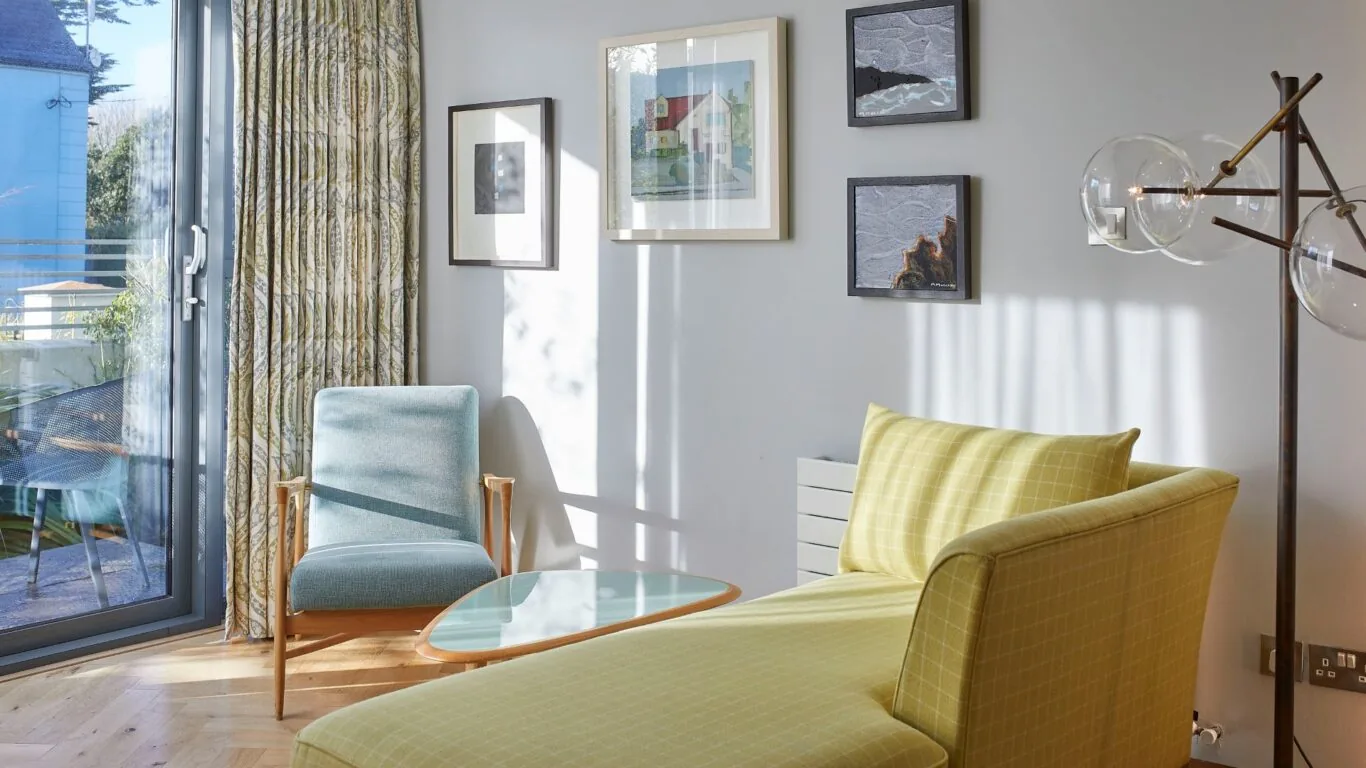 Bright hotel room with stylish seating, artwork, and large window revealing a balcony with lush garden views.