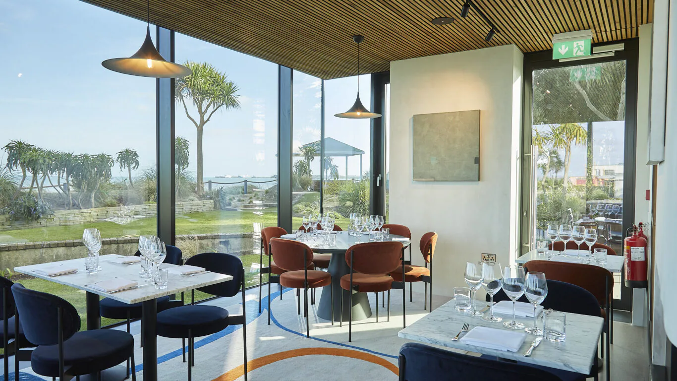 Bright hotel dining room with sea views, stylish seating, modern decor, surrounded by lush gardens. Relaxing dining experience.