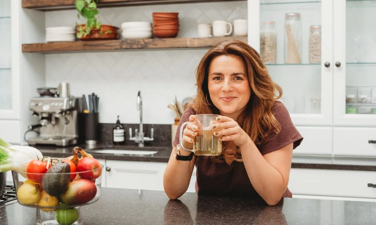 A woman sits at a kitchen counter, holding a mug and smiling. There is a bowl of fruit on the counter and kitchenware on shelves in the background.