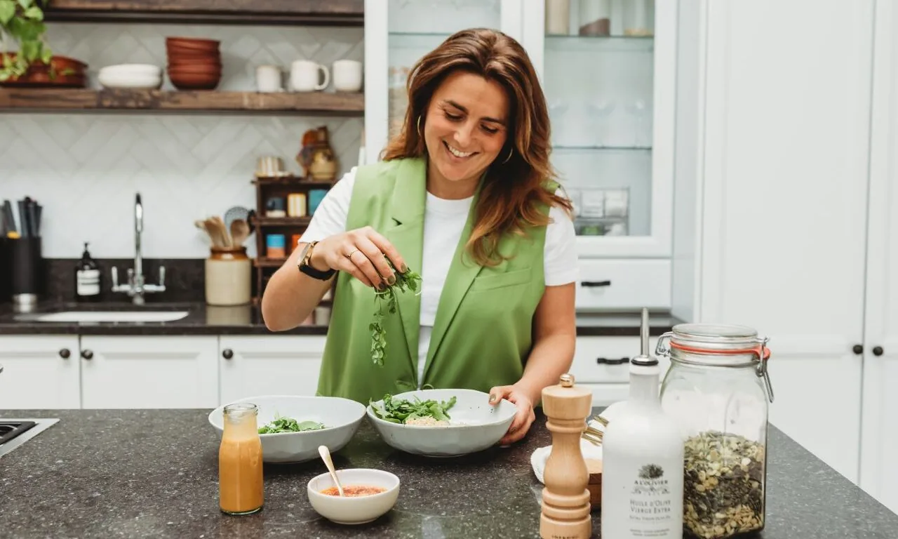 A woman in a green vest sprinkles herbs over a salad in a modern kitchen, with various ingredients and utensils on the counter.