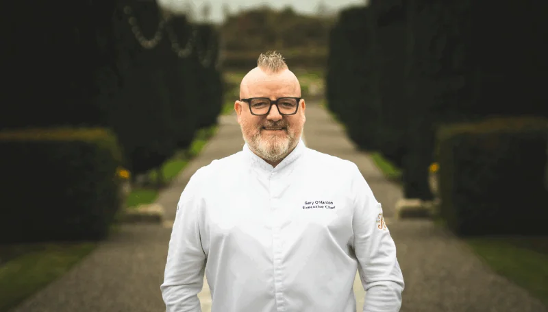 A chef wearing a white jacket stands outdoors on a gravel path lined with trimmed hedges, facing the camera and smiling.