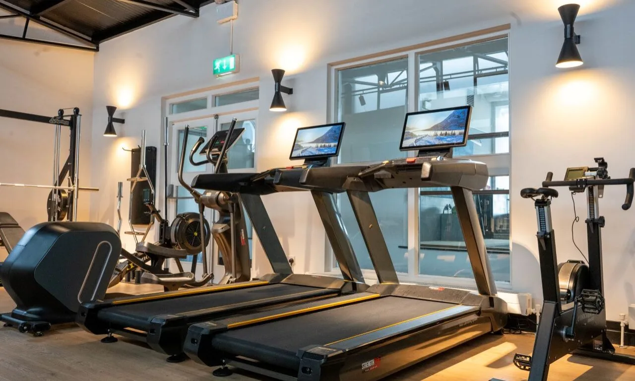 Modern gym with two treadmills, exercise bikes, and elliptical machines, all facing a glass interior wall under ceiling lights.