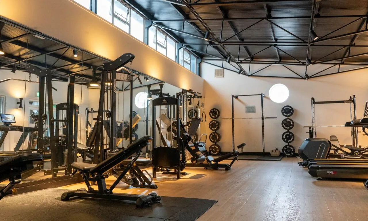 A modern gym with weight machines, benches, free weights, treadmills, mirrors, and wooden flooring under a black metal ceiling.