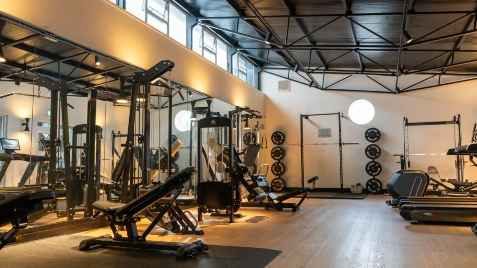 A modern gym with weight machines, benches, free weights, treadmills, mirrors, and wooden flooring under a black metal ceiling.