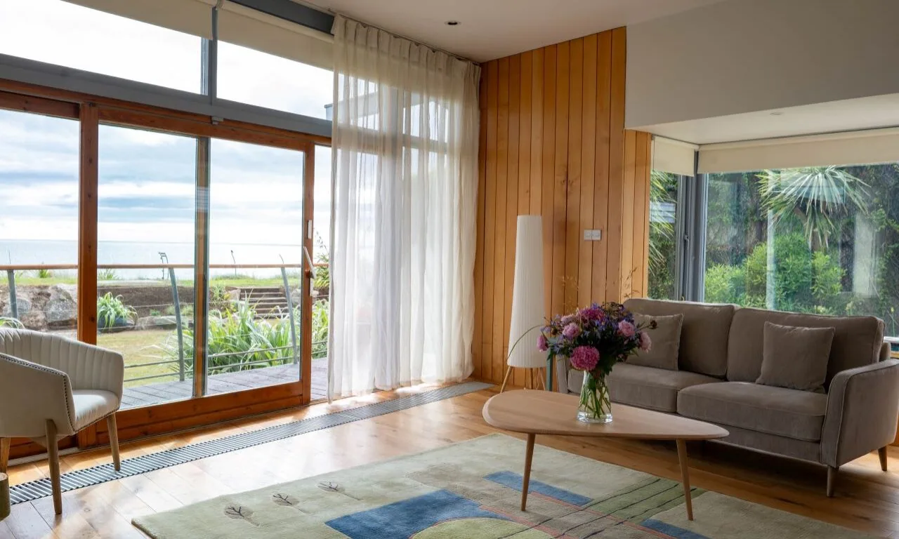 Modern living room with large windows, a gray sofa, a wooden coffee table with a flower vase, a light armchair, and a view of a garden and the sea outside.