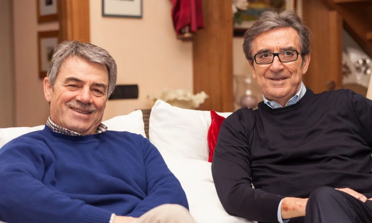 Two older men sit on a white couch indoors, smiling at the camera. Wooden furniture and framed pictures are visible in the background.