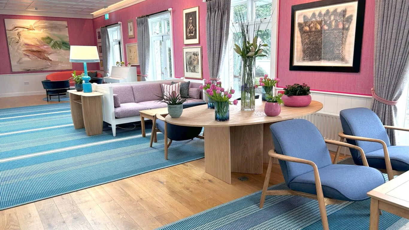 Spacious hotel lounge with pink walls, blue chairs, vibrant art, and fresh flowers, creating a cosy, inviting atmosphere.